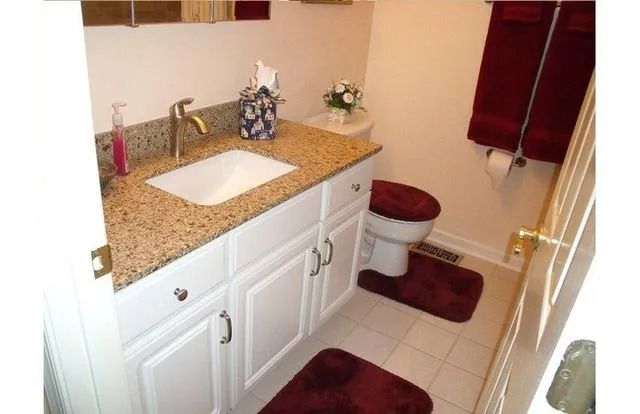 Small bathroom with white vanity, speckled countertop, toilet, burgundy rugs, and red window curtains.