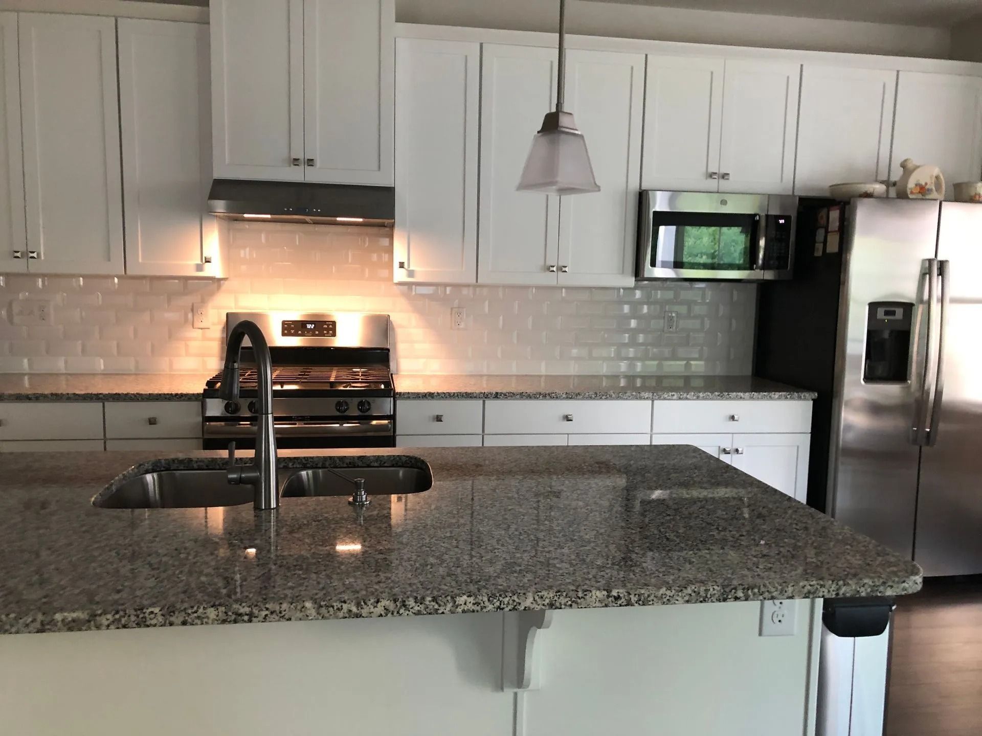 Modern kitchen with white cabinets, granite island, stainless stove, fridge, microwave, and pendant light.