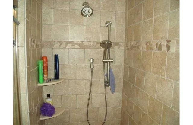 Bathroom shower with beige tile, handheld showerhead, and wall shelf holding toiletries.