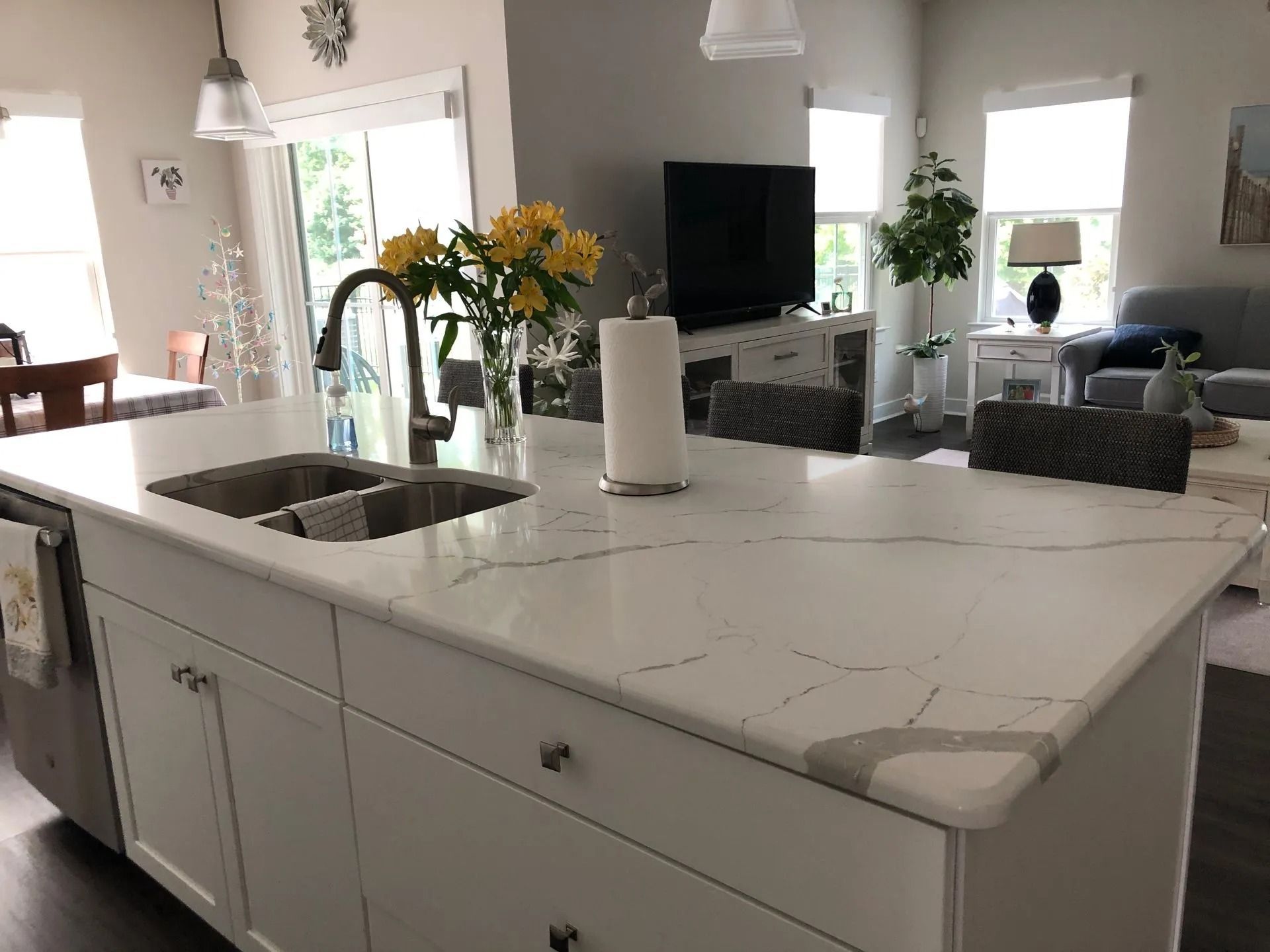 Bright kitchen island with marble countertop, sink, white cabinets, and flowers in a sunlit open living room
