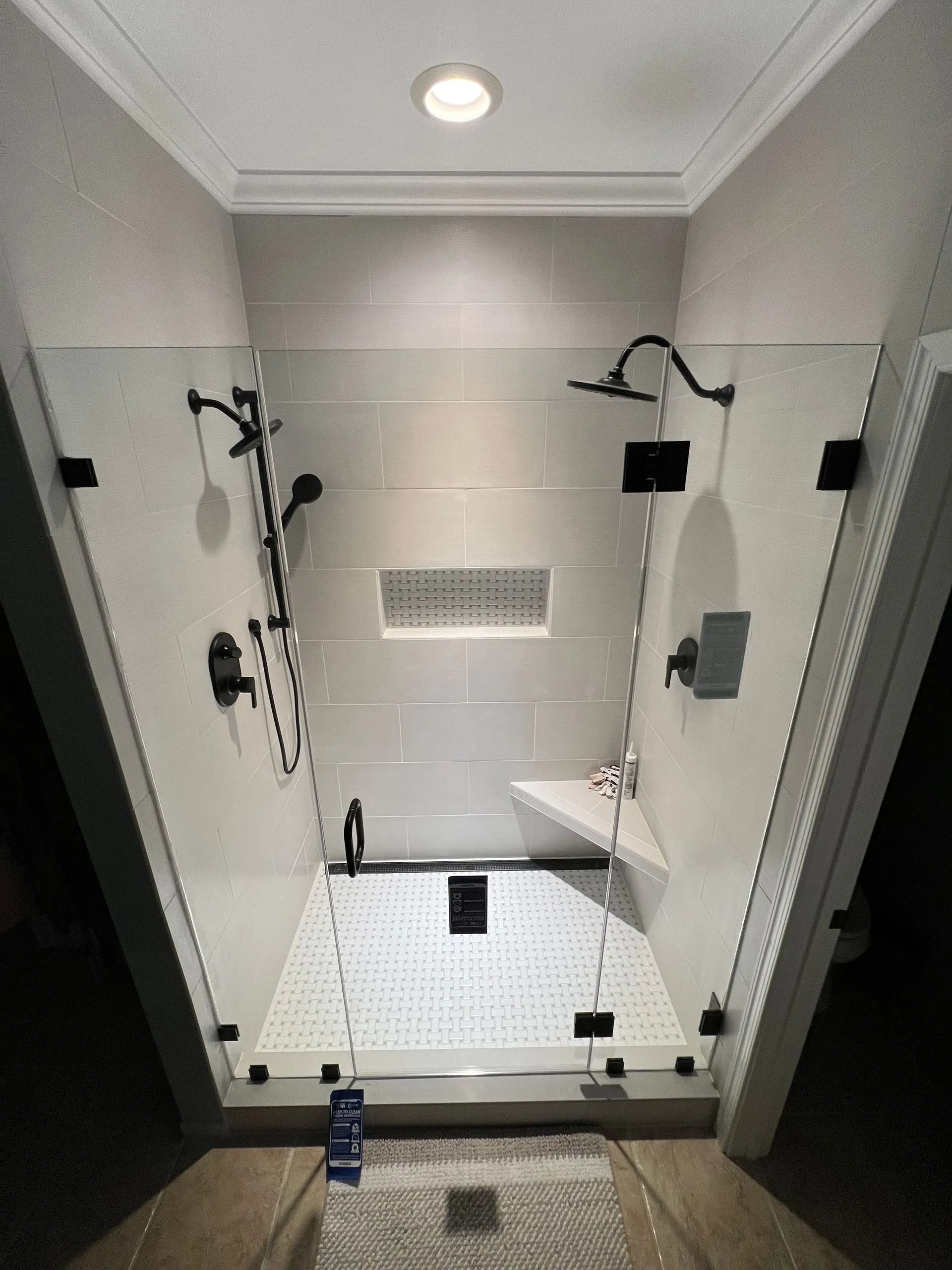 Empty tiled shower stall with handheld showerheads, drain, and recessed light in a bathroom.