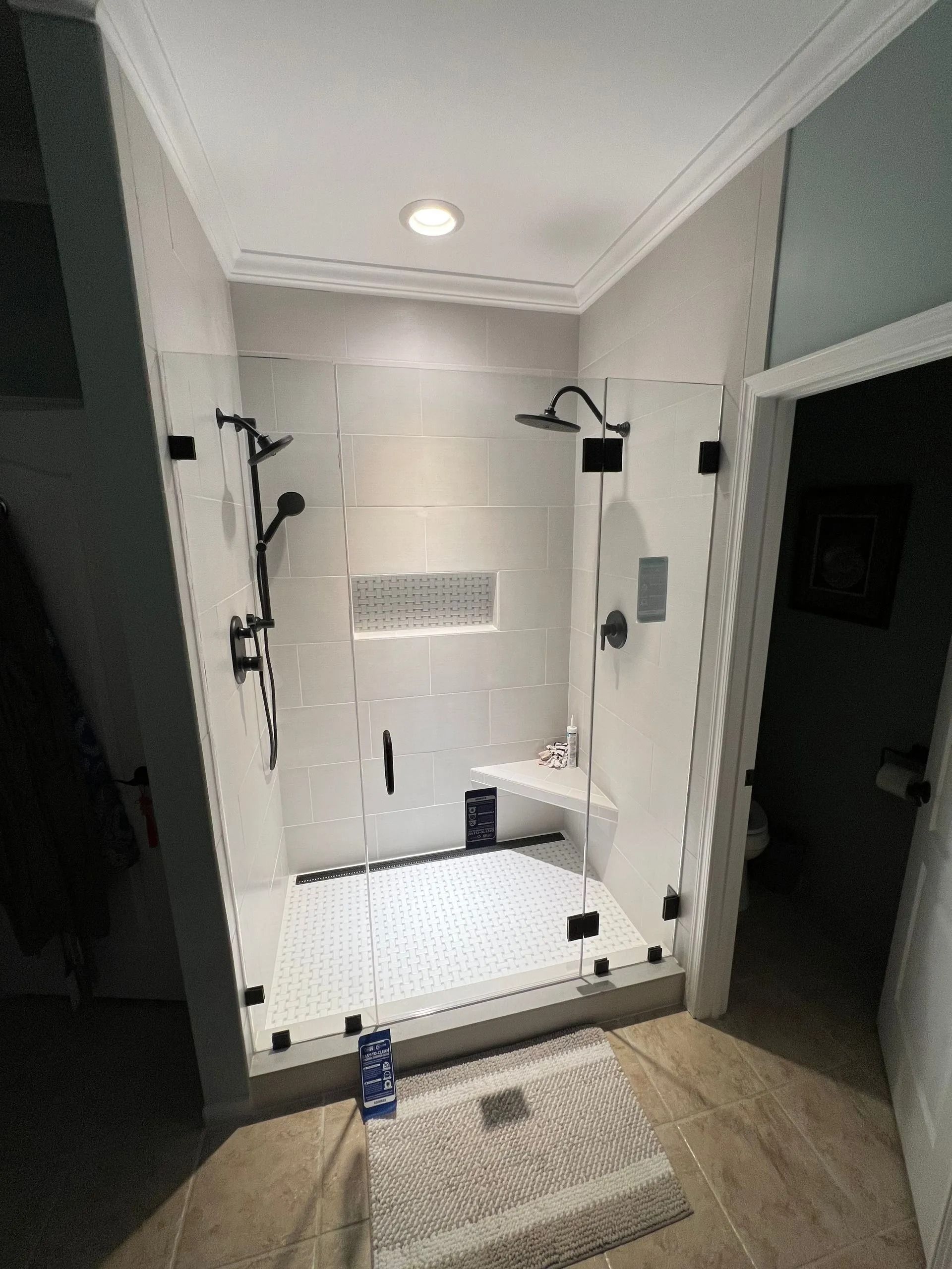 Walk-in shower with white tile, black fixtures, and a bath mat in a bathroom doorway