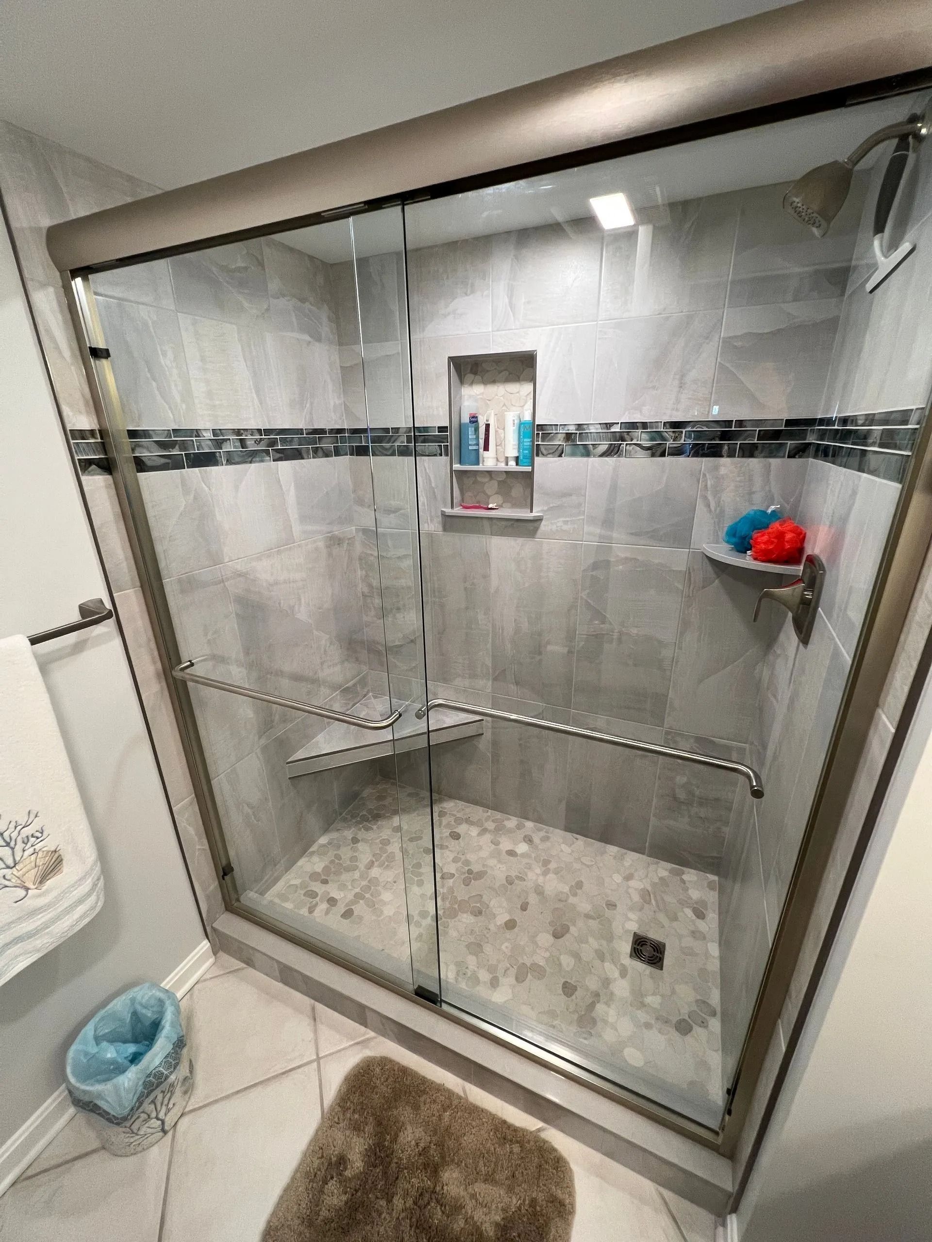 Glass walk-in shower with gray tile, chrome trim, corner shelves, and a shower mat outside the door