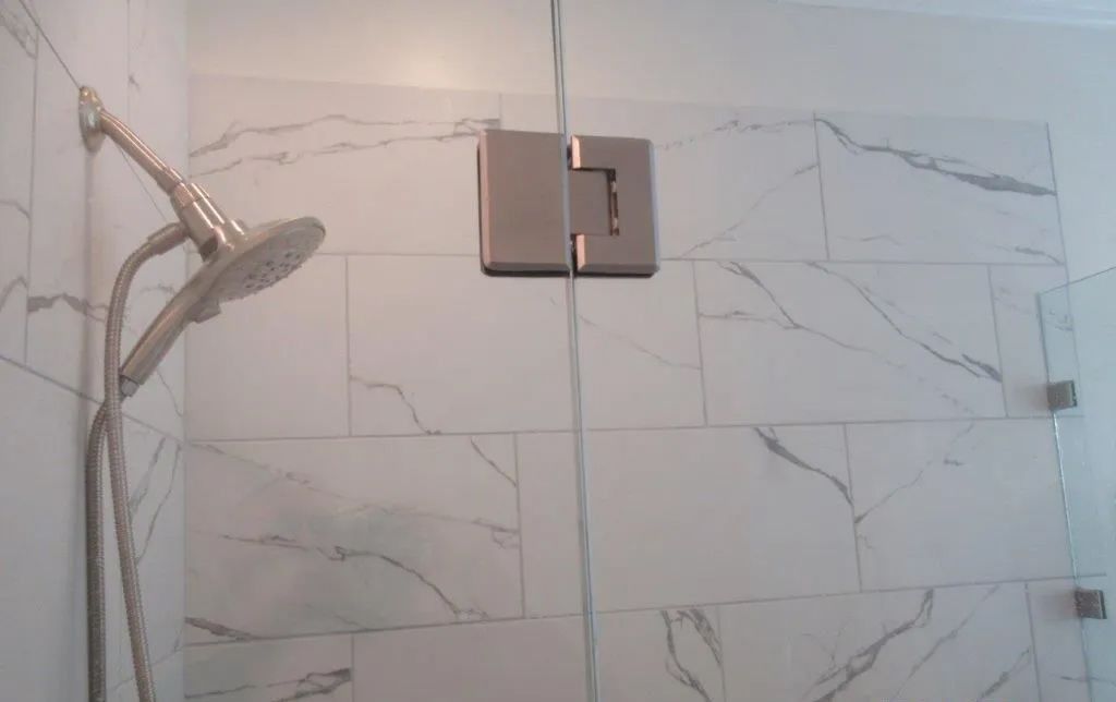 Bathroom shower with marble-look tile walls, glass door hinges, and a chrome showerhead on the left