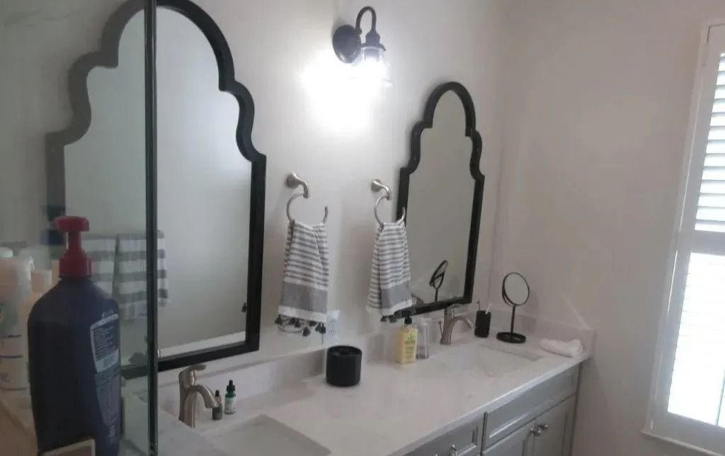 Bathroom vanity with two ornate mirrors, white countertop, soap dispensers, and a window with blinds.