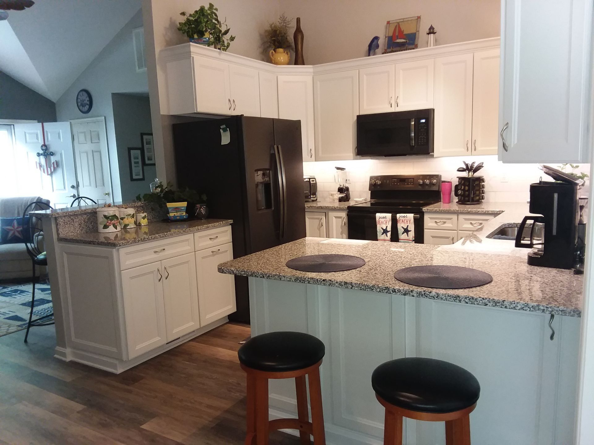 Bright kitchen with white cabinets, granite counters, black appliances, and two stools at an island