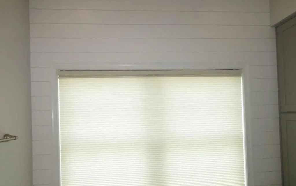 Large window with closed white blinds in a light wood-paneled room