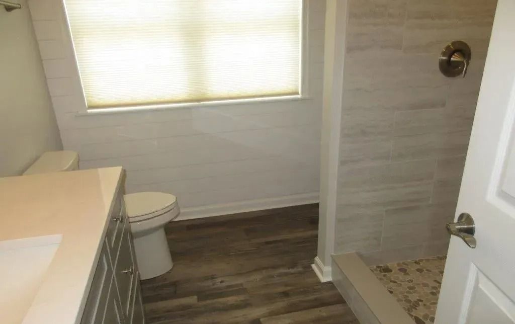Small bathroom with toilet, window, wood-look floor, and walk-in shower visible through open door