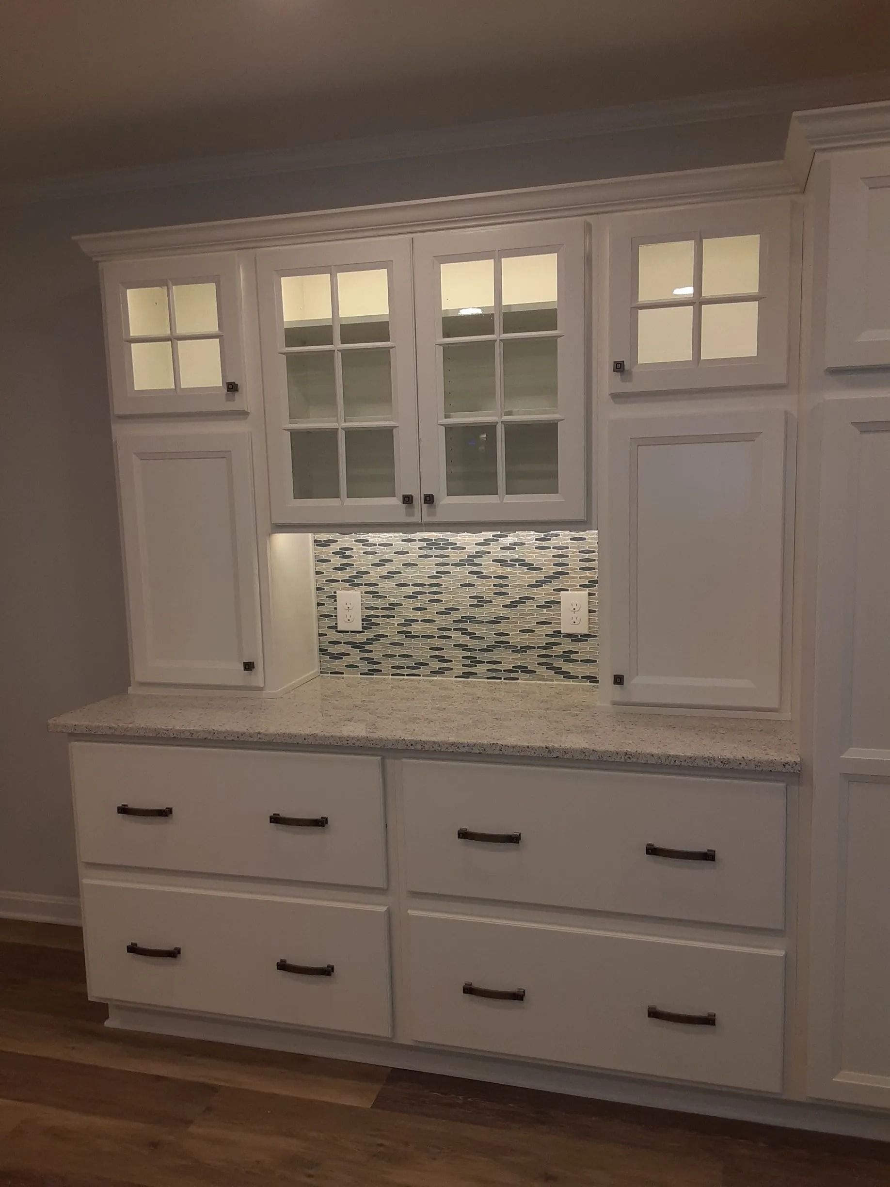 White built-in kitchen cabinet with glass doors, mosaic backsplash, and drawers beneath a granite countertop.