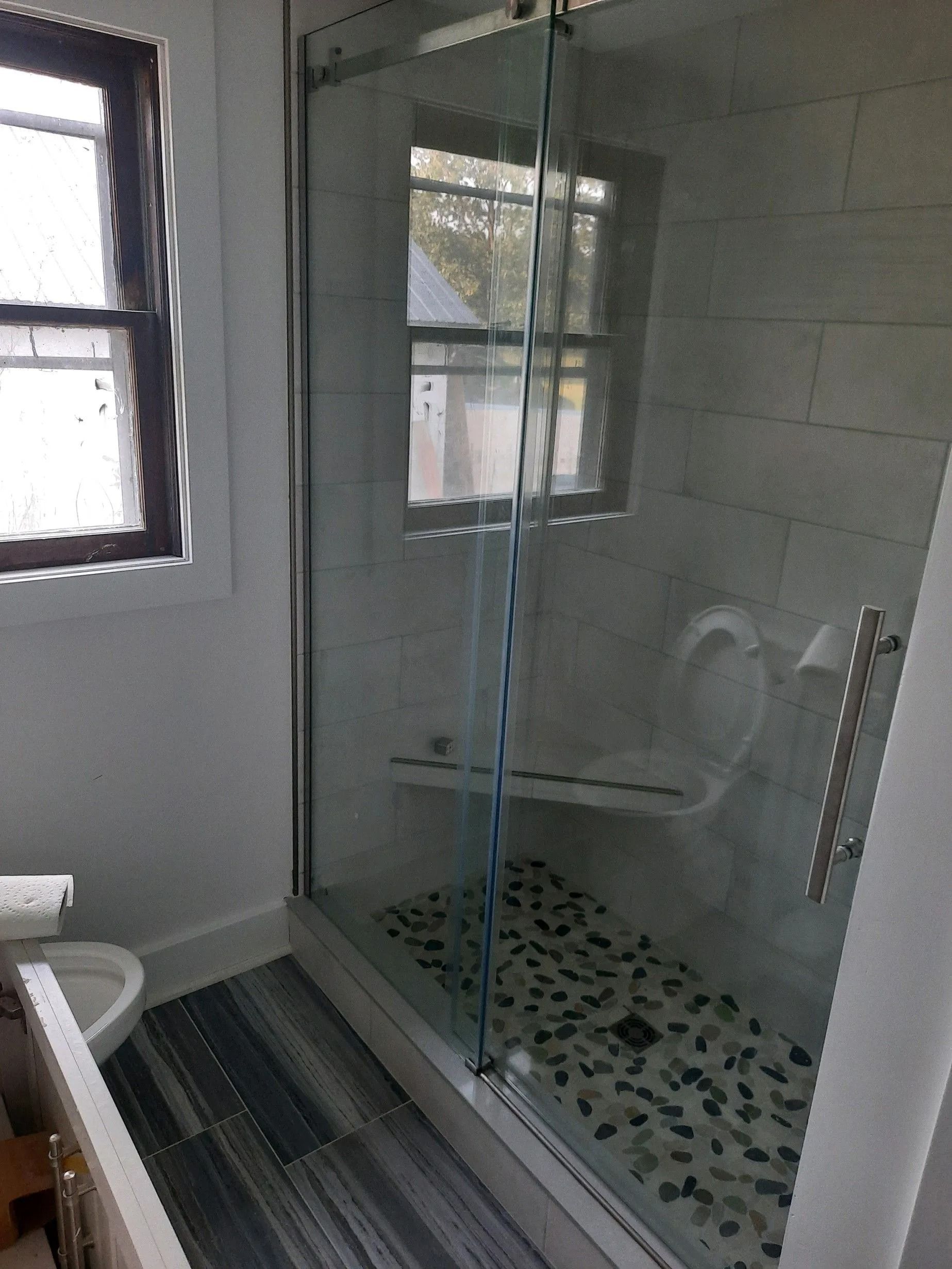 Small bathroom with a glass shower enclosure, pebble tile floor, and a window beside the toilet.