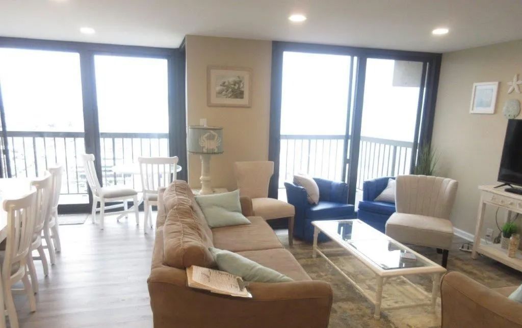 Bright living room with tan and blue sofas, glass coffee table, and balcony doors with city views.