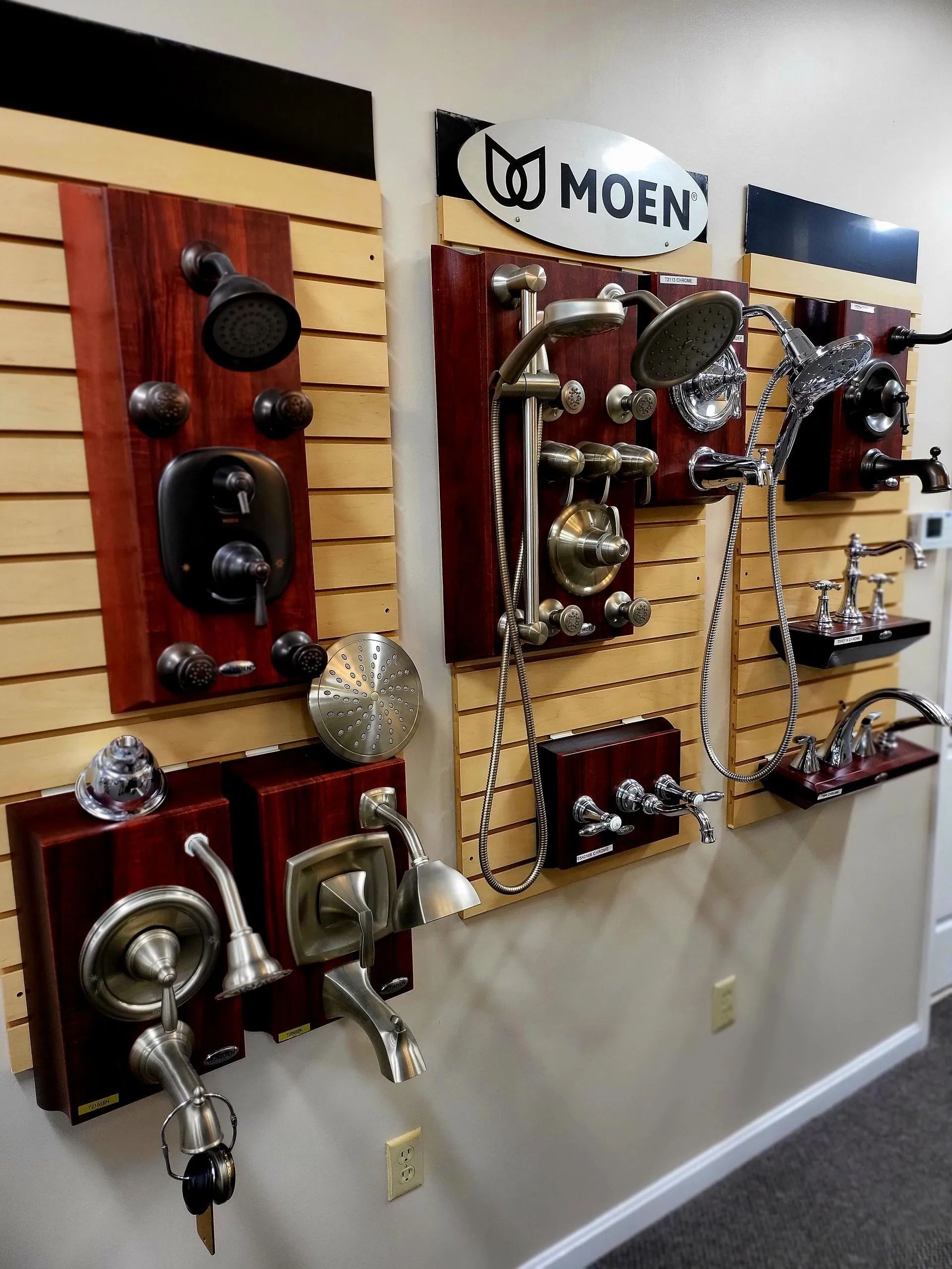 Moen wall display of assorted chrome and bronze bathroom faucets and shower fixtures on wood panels