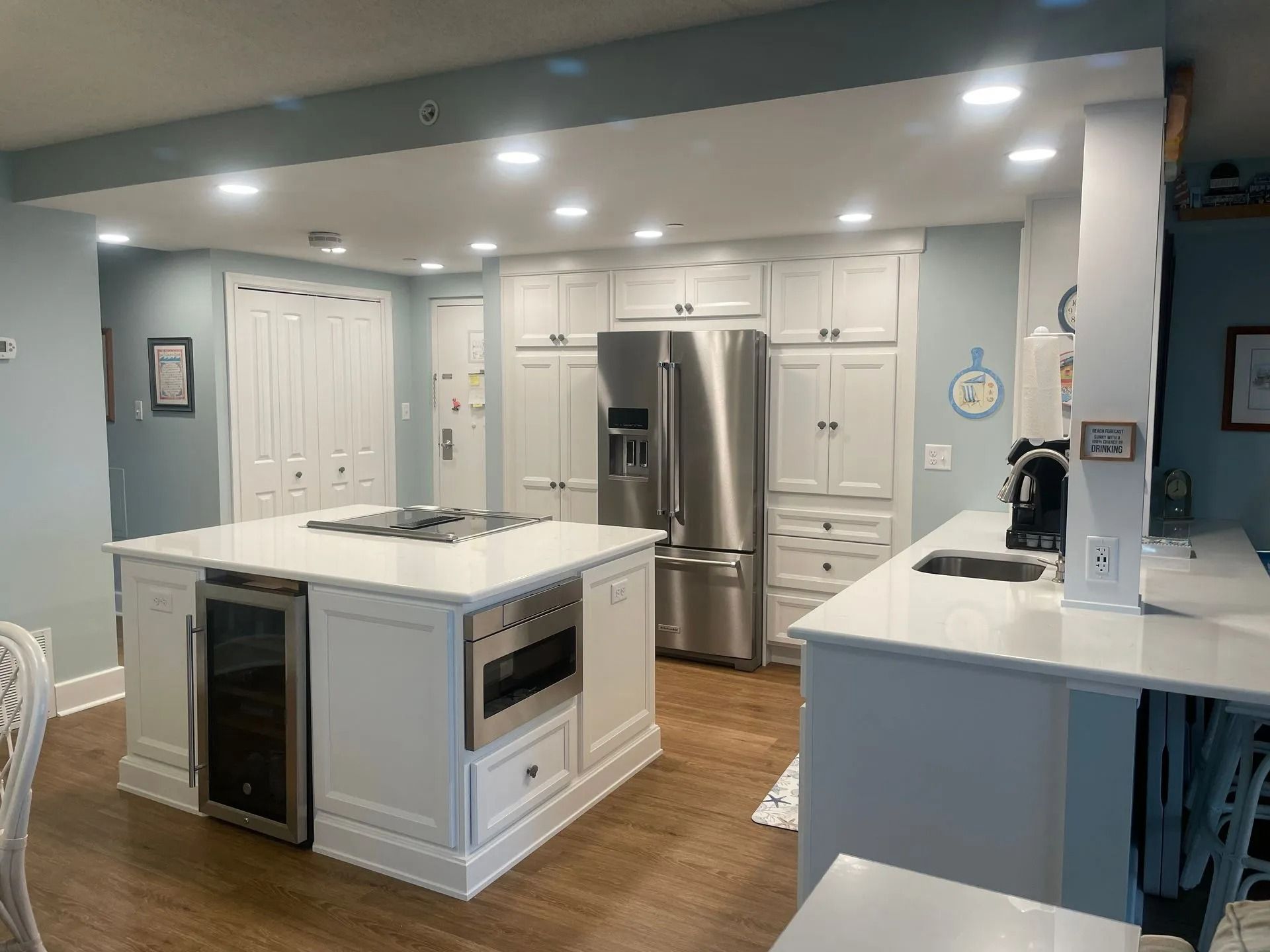 Modern white kitchen with island, stainless steel fridge, and built-in oven, seen in bright recessed lighting