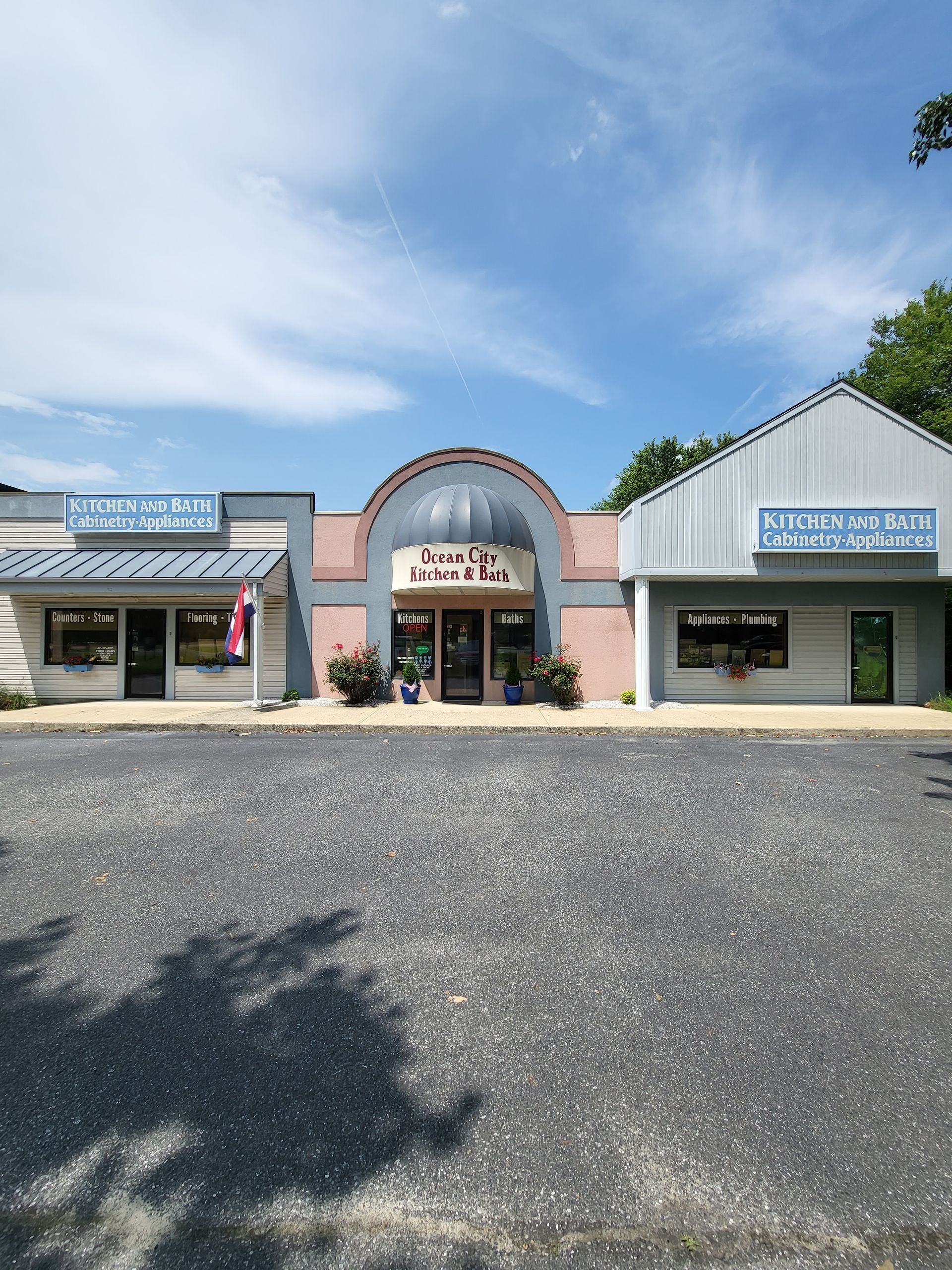 Small row of storefronts with a central arched entrance, blue signs, and a gravel parking lot under a clear sky