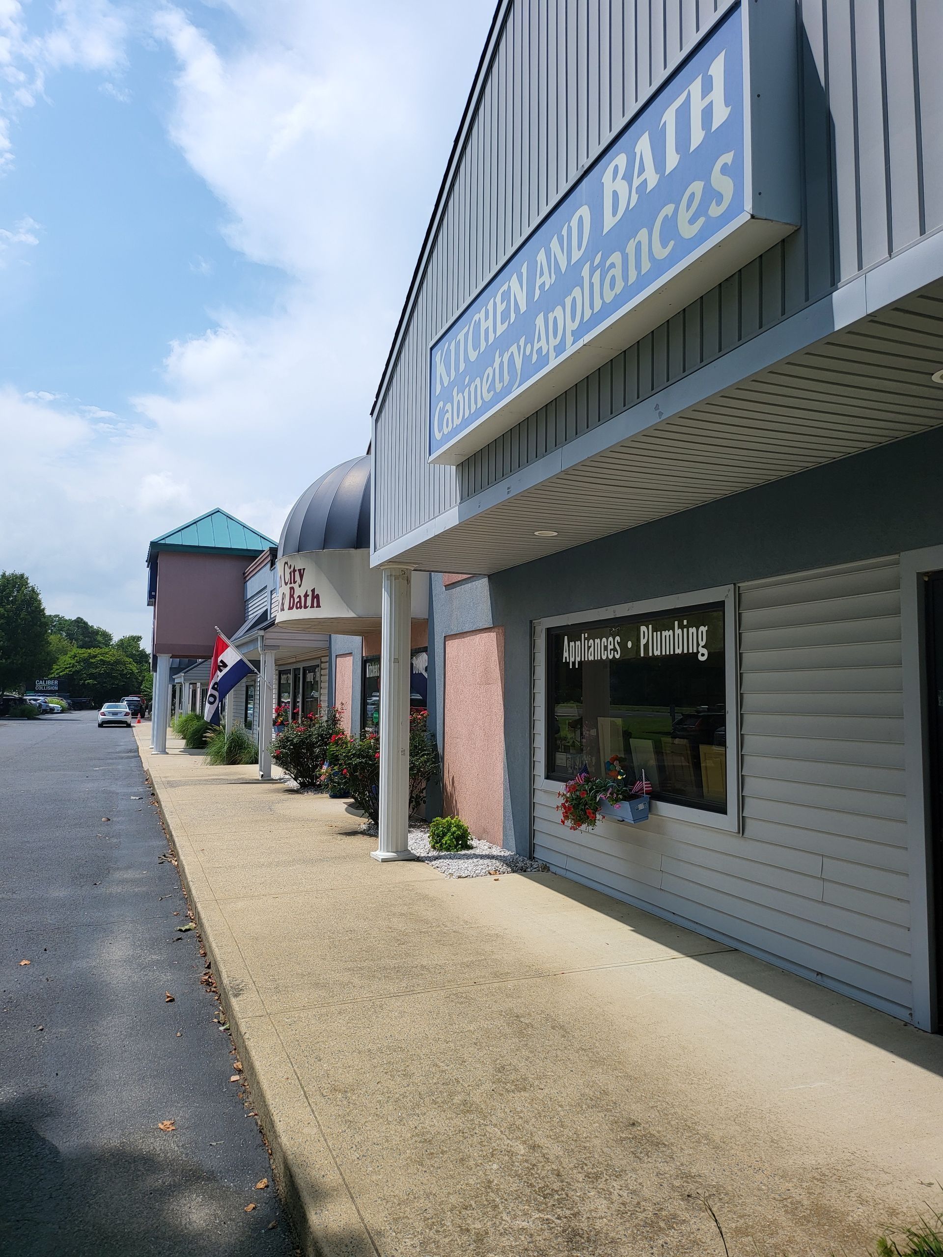 Strip mall storefronts with a blue sign, gravel walkway, and parking lot on a sunny day