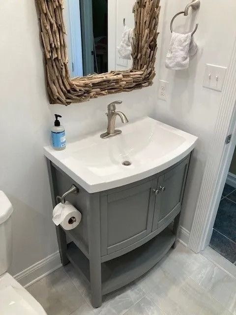 Small gray bathroom vanity with white sink, mirror, soap dispenser, and toilet paper holder beside a shower