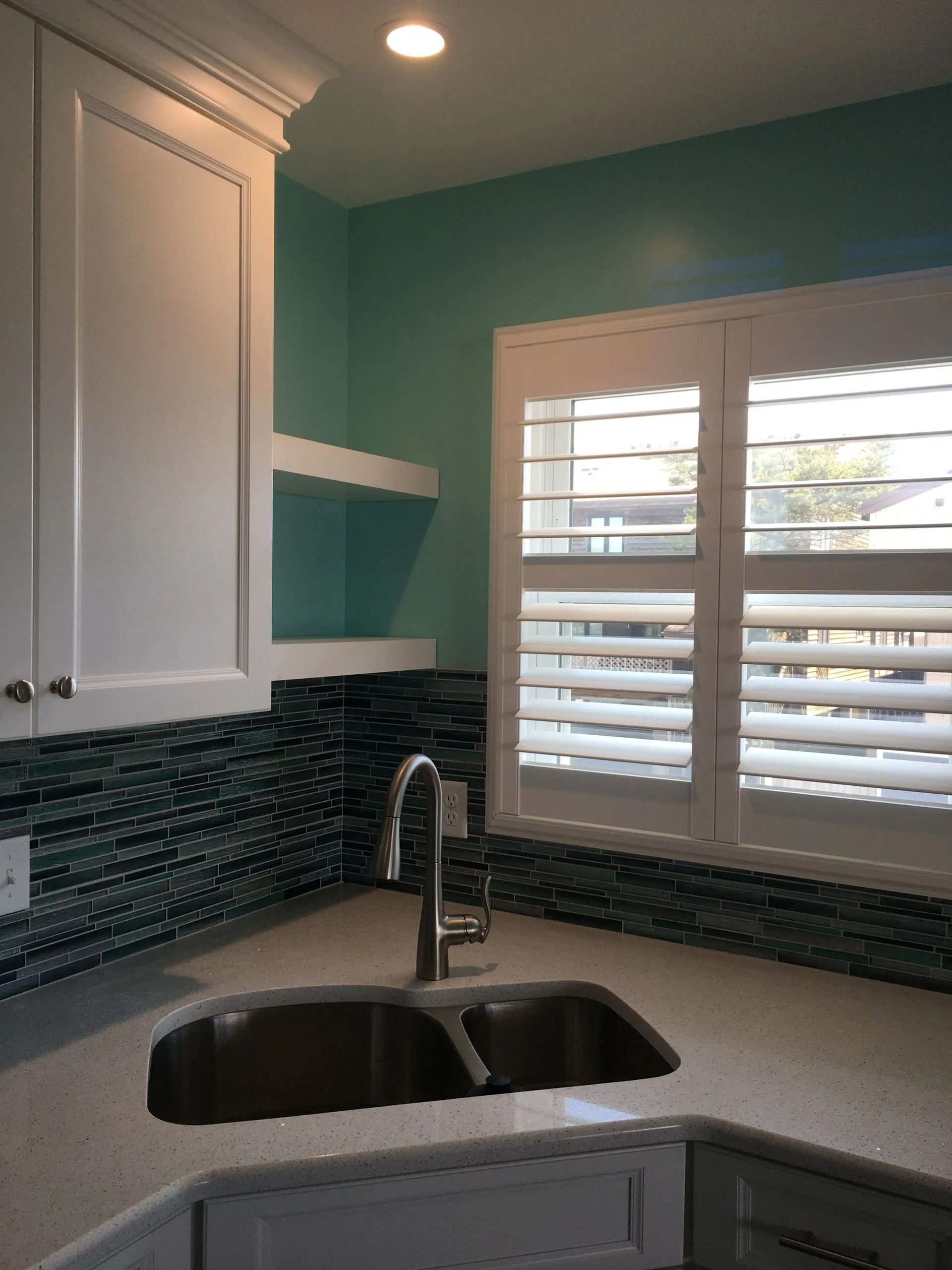 Bright kitchen corner with white cabinets, black sink, teal walls, and window shutters.