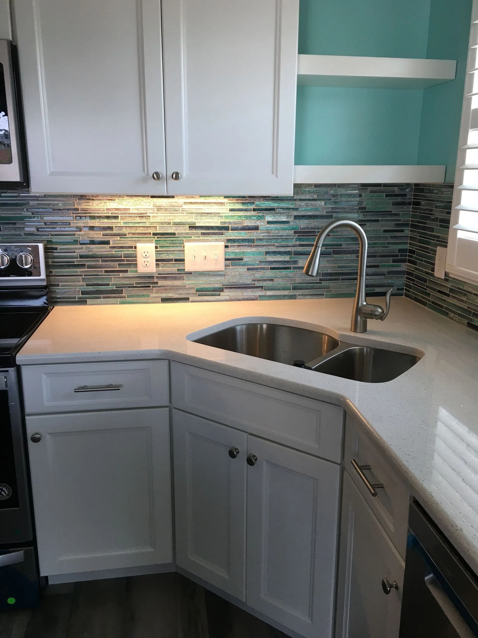 Compact kitchen corner with white cabinets, teal mosaic backsplash, double sink, and chrome faucet