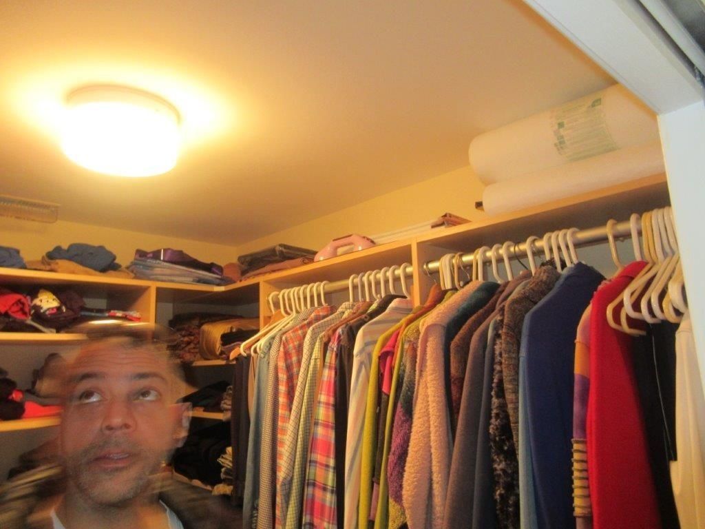 Man in a closet room beside hanging clothes and shelves, lit by a ceiling light.