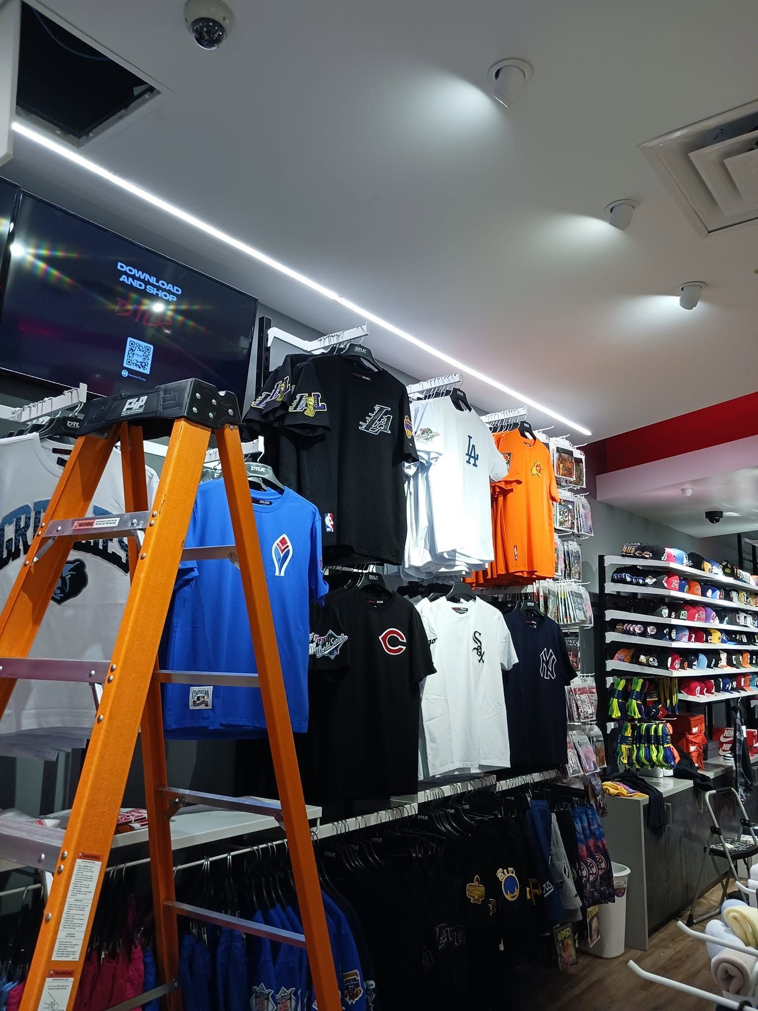 Orange ladder in a clothing store aisle with racks of shirts and jackets under bright ceiling lights