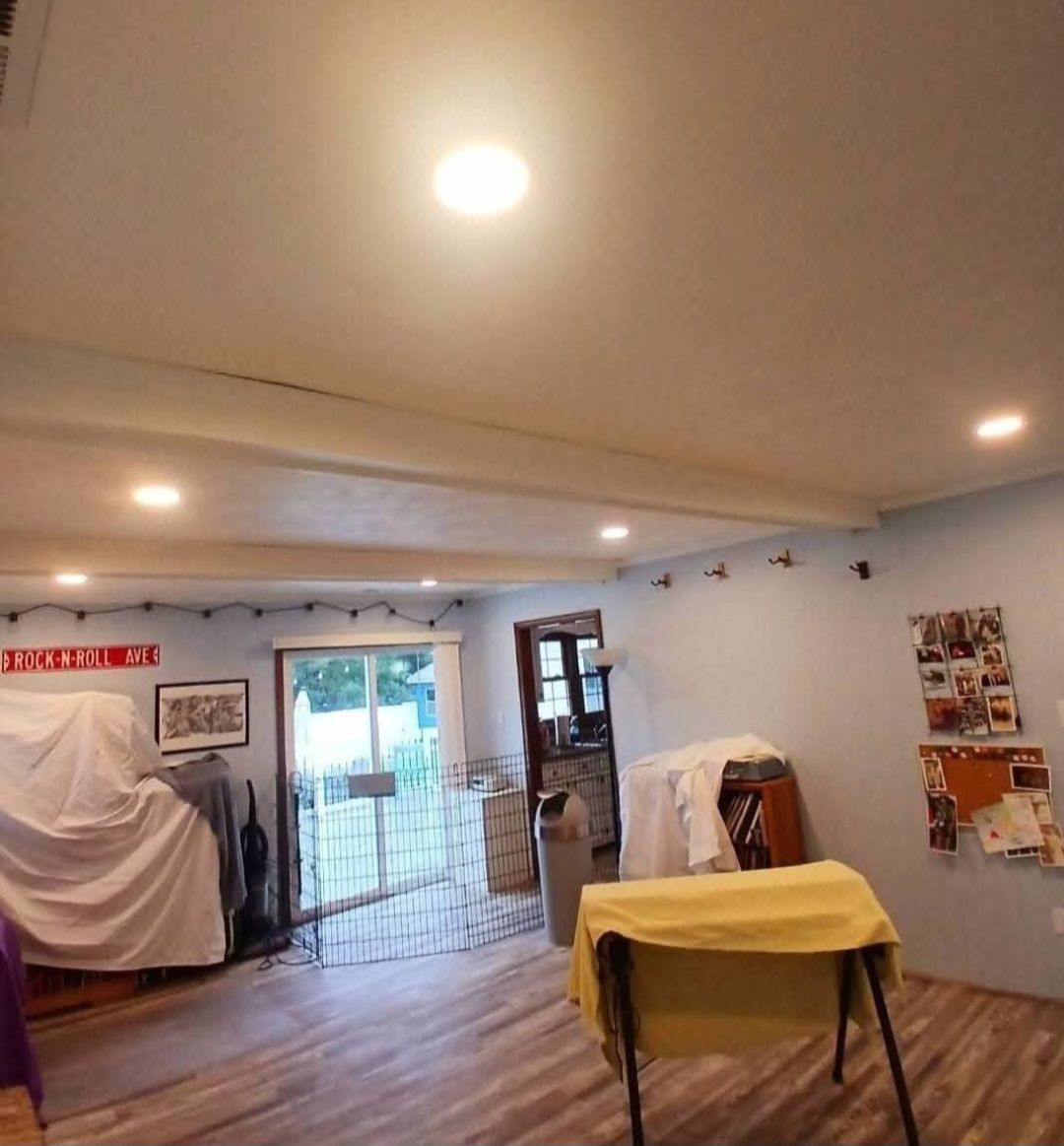 Spacious room with wood floor, ceiling lights, covered furniture, and a table draped in yellow cloth by the window