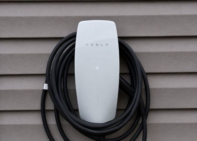 White wall-mounted electric vehicle charger with coiled black cable against gray siding