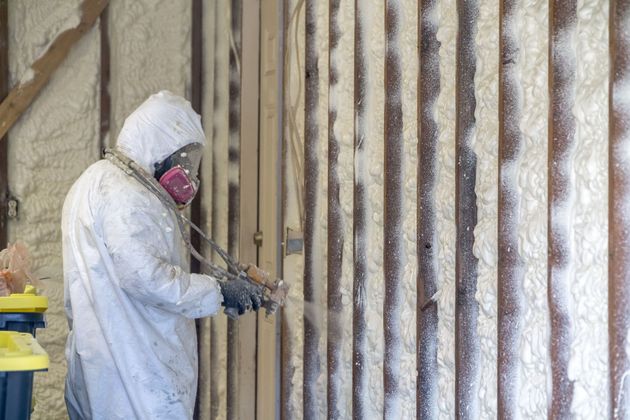 Worker in white protective suit and respirator spraying insulation foam inside a framed wall.