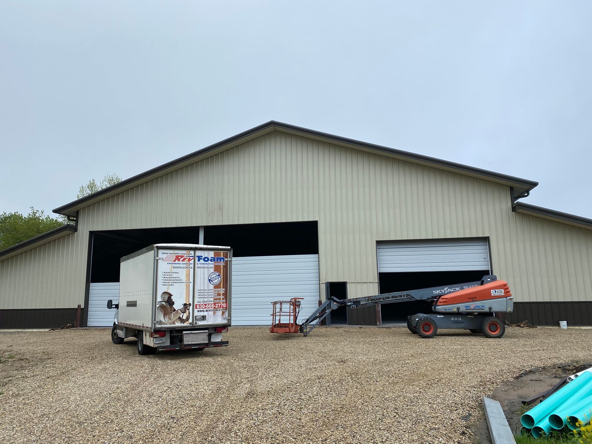 Large beige barn with open doors, farm equipment parked outside on a gravel lot.