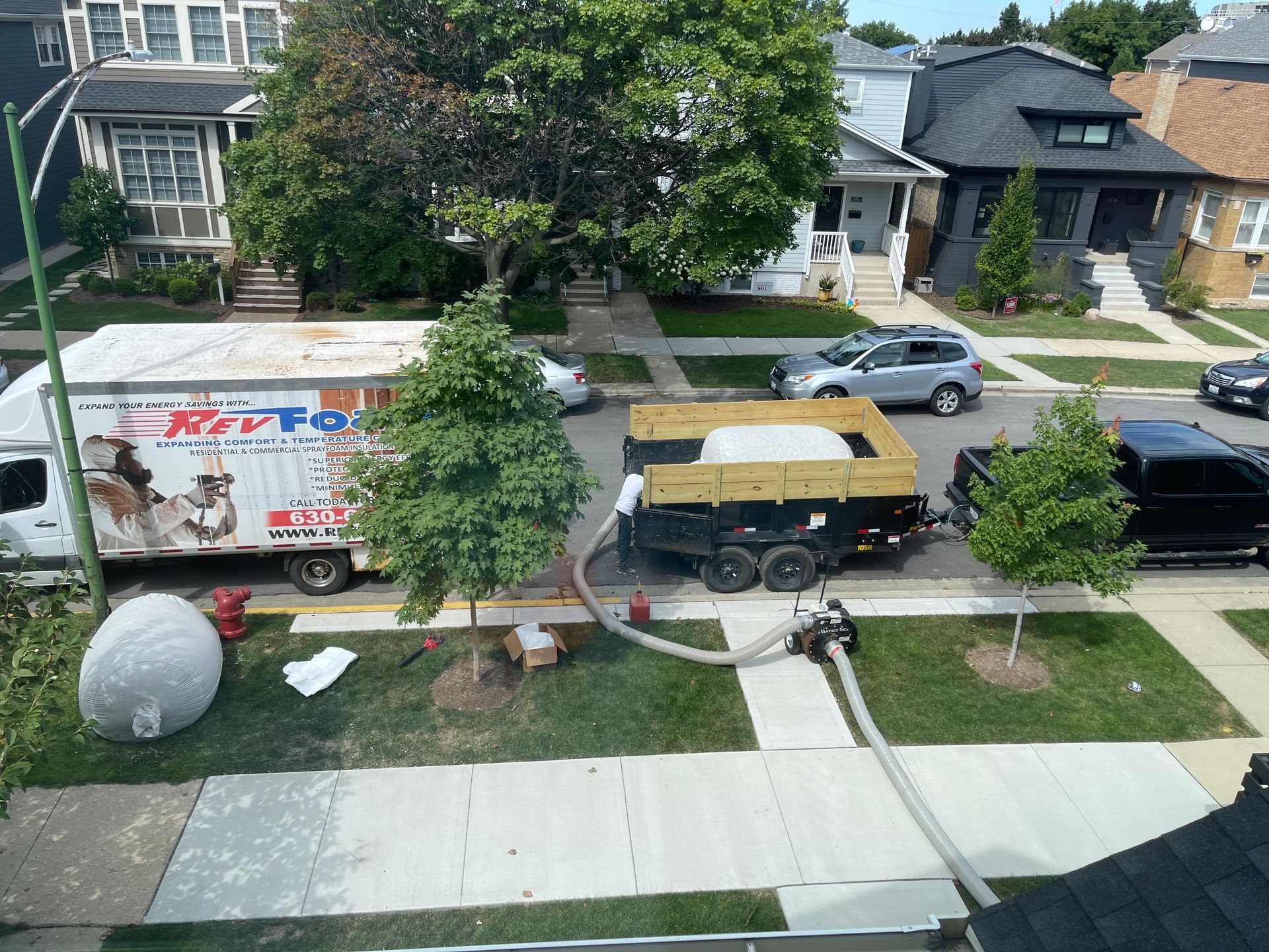 Street view of a parked yellow-and-black van, box truck, and car along a residential sidewalk with hoses across the lawn
