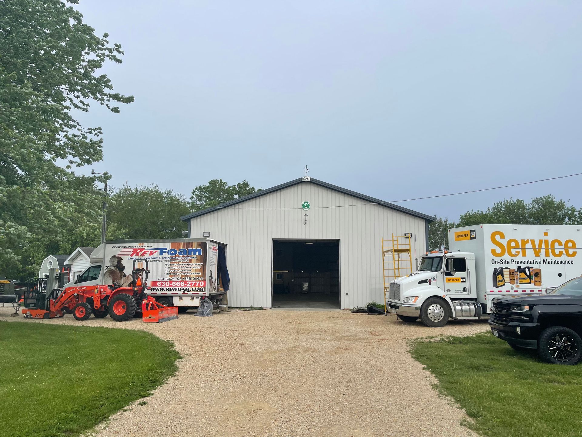 Service trucks and a tractor parked outside a white metal equipment building on a gravel lot