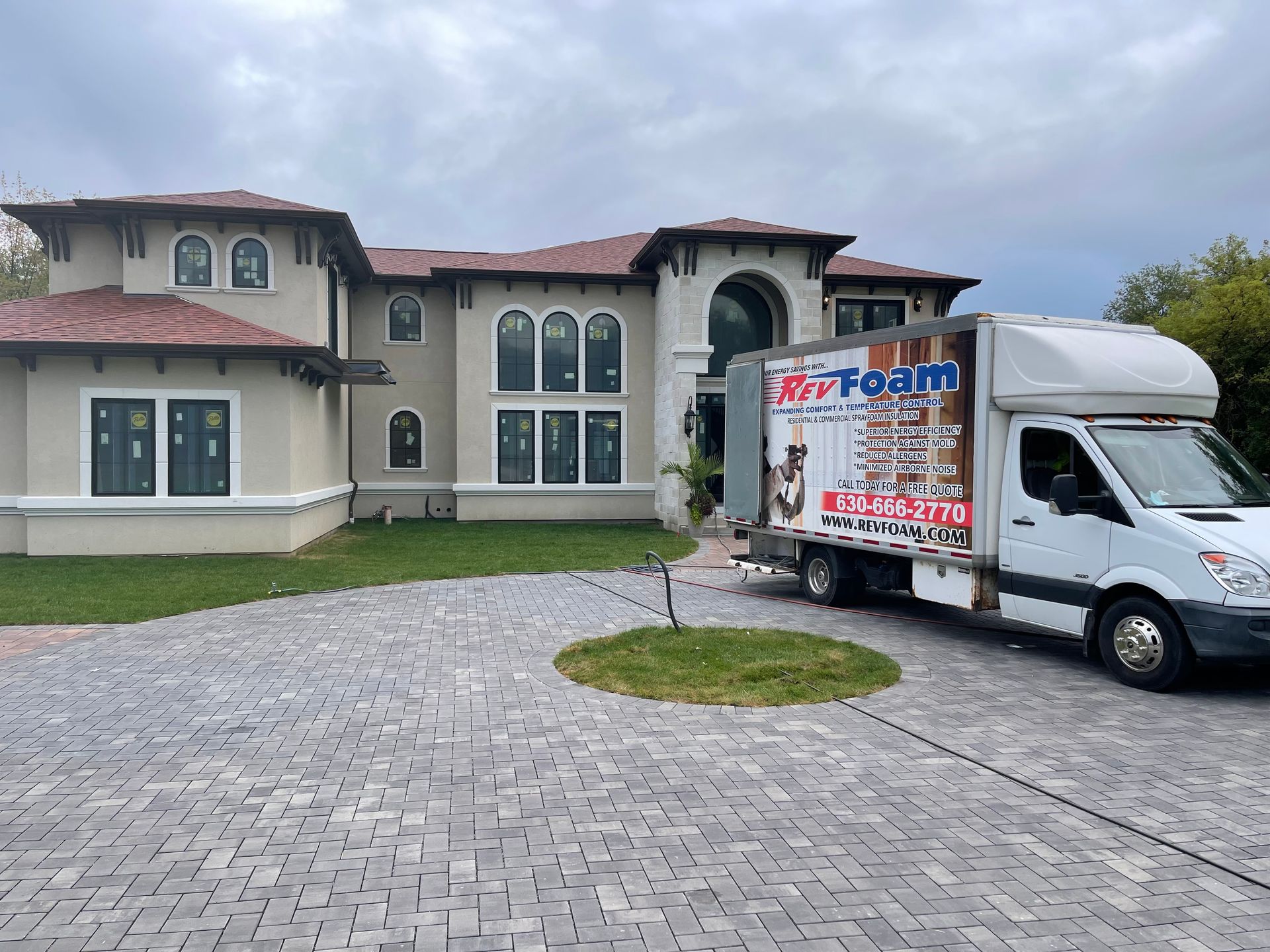 Moving truck parked in front of a large cream-colored house with a brick driveway under cloudy skies