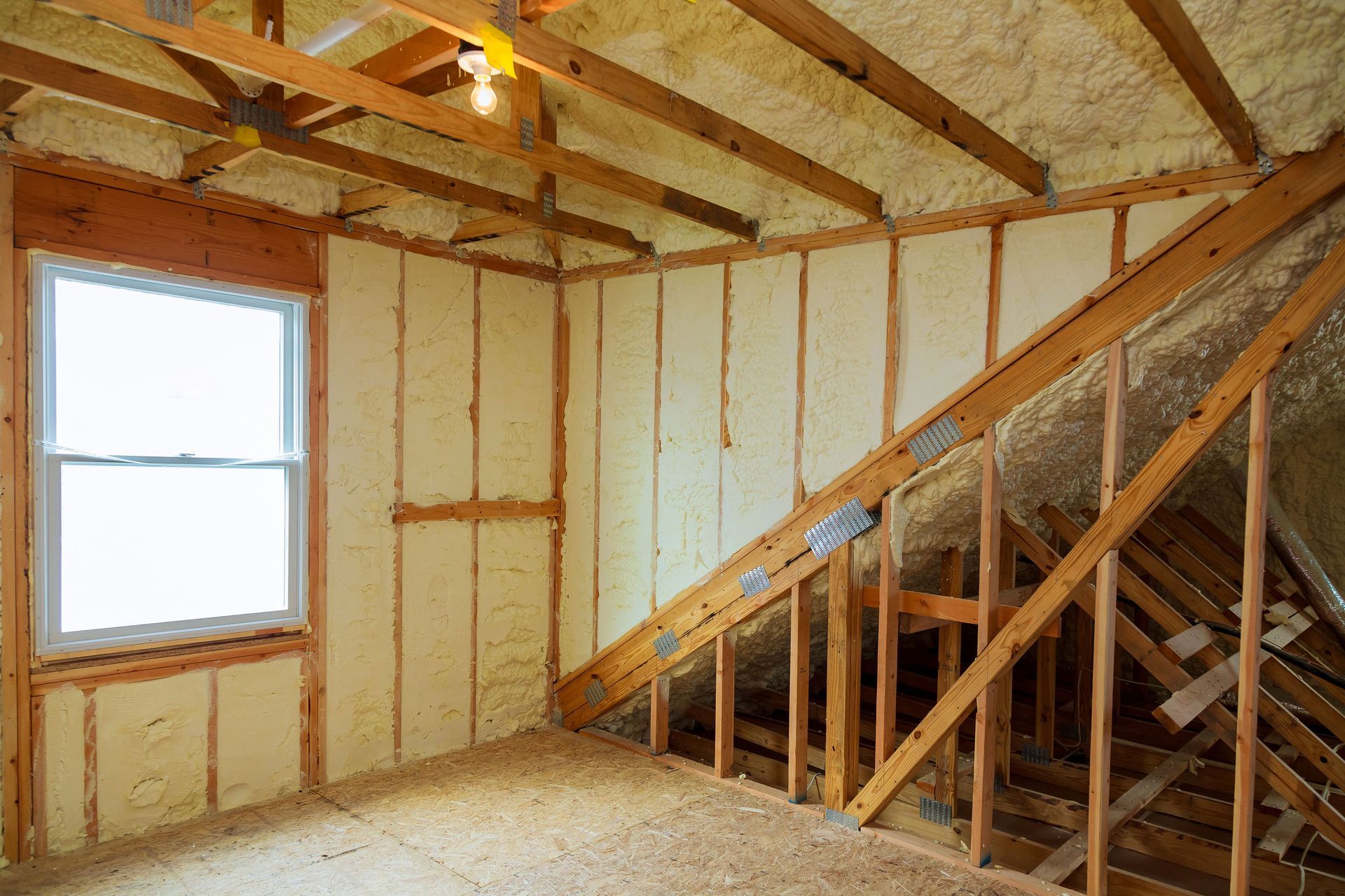 Interior of unfinished attic with exposed wooden framing, insulation, sloped roof, and a window on the left.