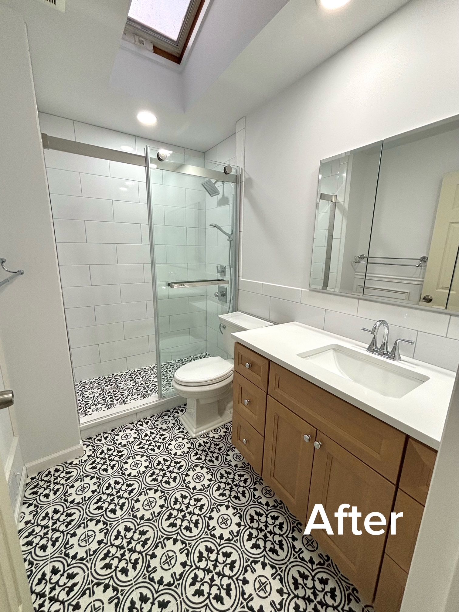 Bright white bathroom with patterned black-and-white tile, wood vanity, mirror, toilet, and glass shower.