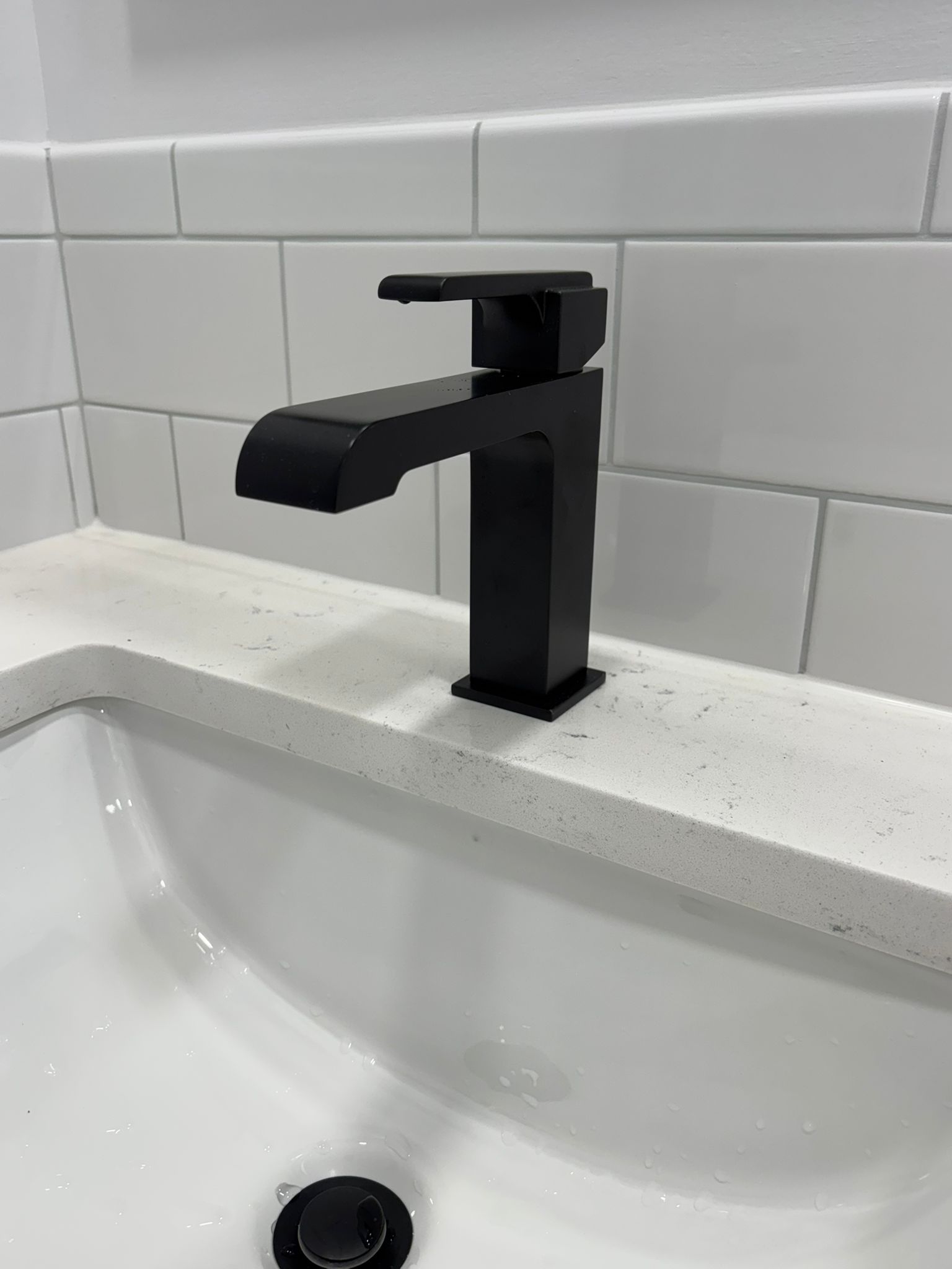 Black faucet on white countertop above a white sink, set against white tiled wall.