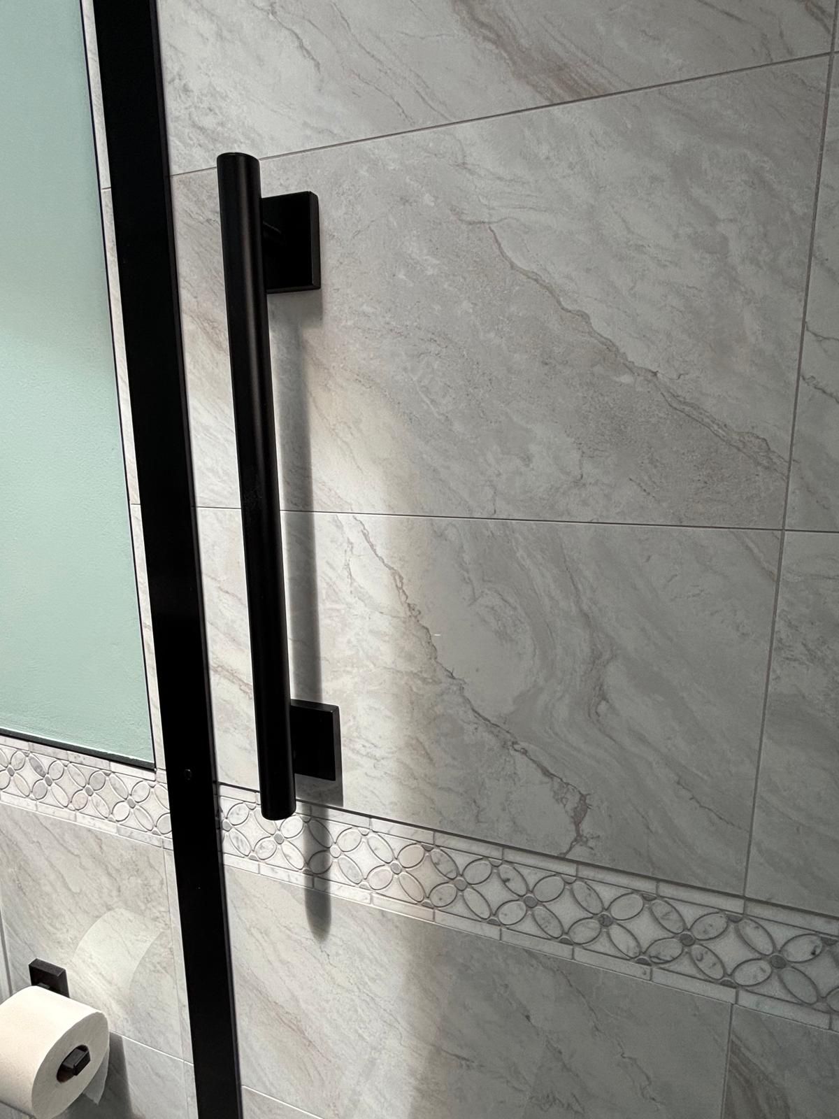Black shower door handle on gray tiled wall. Toilet paper roll is visible in the lower left corner.