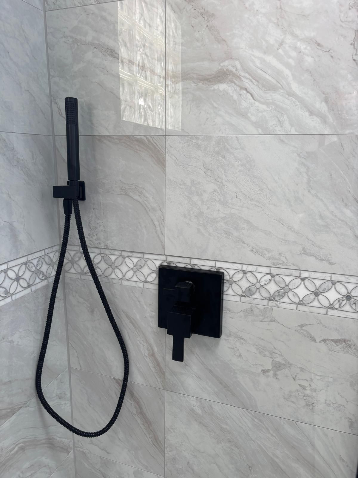 Black shower fixtures against a light gray marbled tile wall. A hand-held shower head hangs on a black hose.