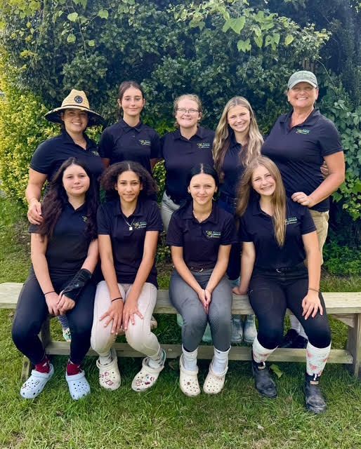 Nine people in dark shirts pose outdoors in two rows in front of a leafy hedge.