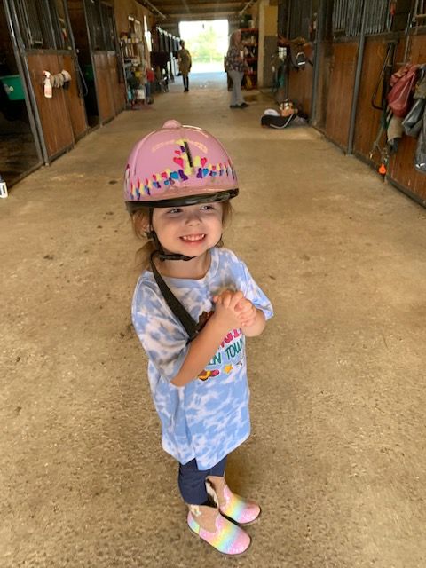 Smiling child wearing a pink helmet, standing in a barn aisle in colorful clothes and shoes
