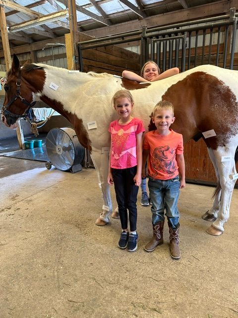 Two children standing beside a brown-and-white horse in a barn, with a smiling woman peeking over the horse.