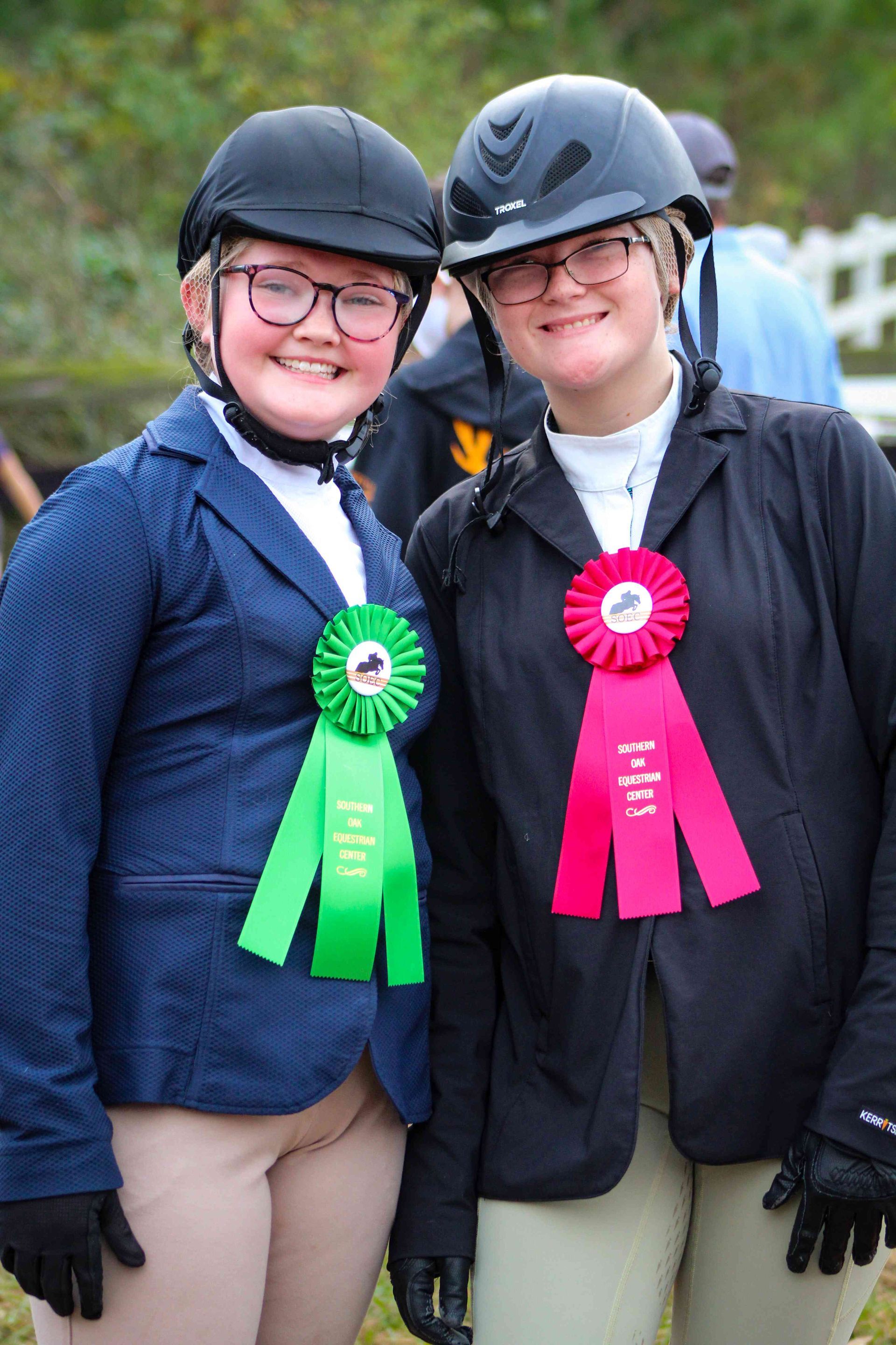 Two riders in riding helmets and show jackets, smiling with green and pink ribbons.