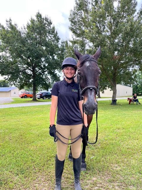 Person standing with a horse in a grassy field, wearing riding gear and smiling at the camera.