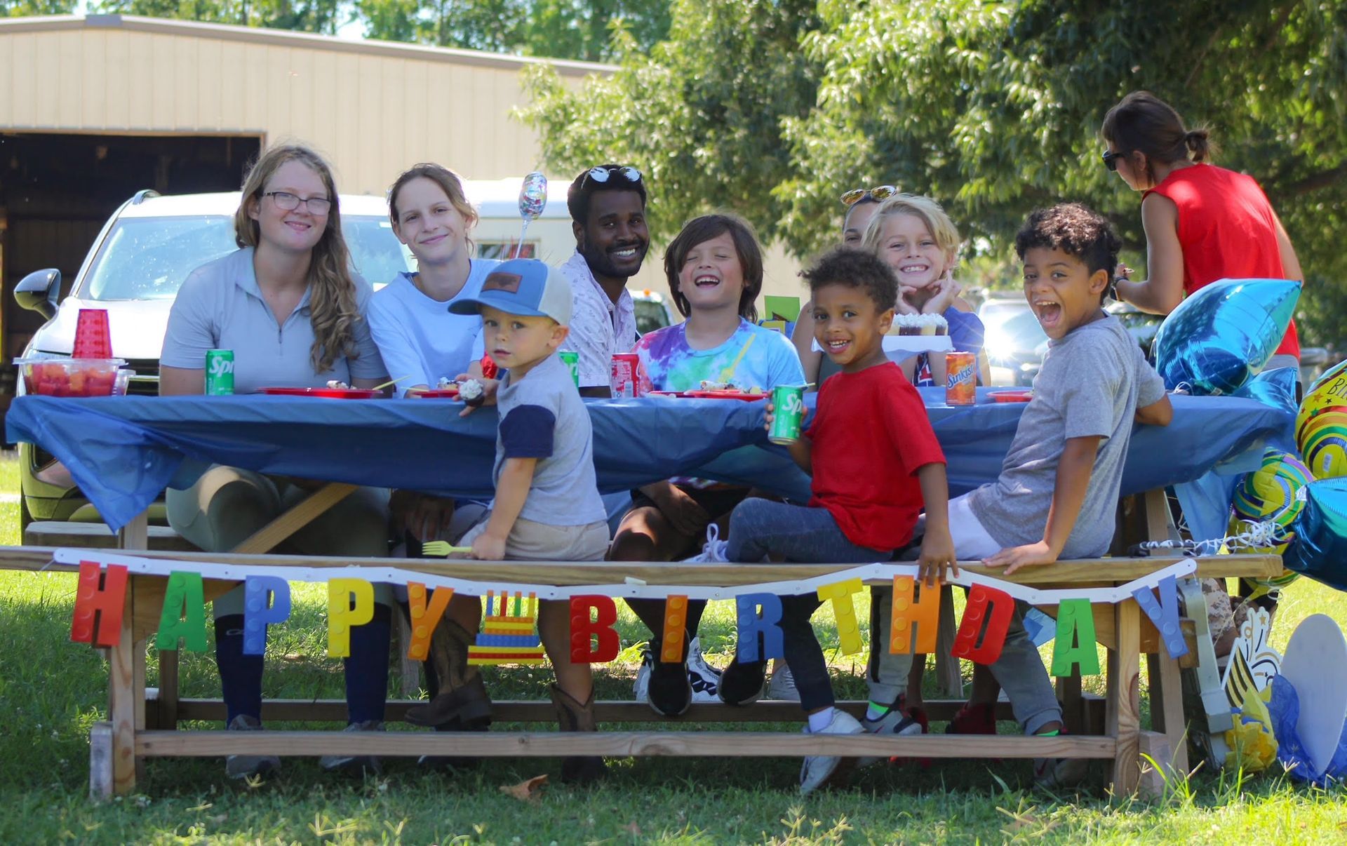 People seated at a colorful outdoor birthday party table with “Happy Birthday” banner and balloons