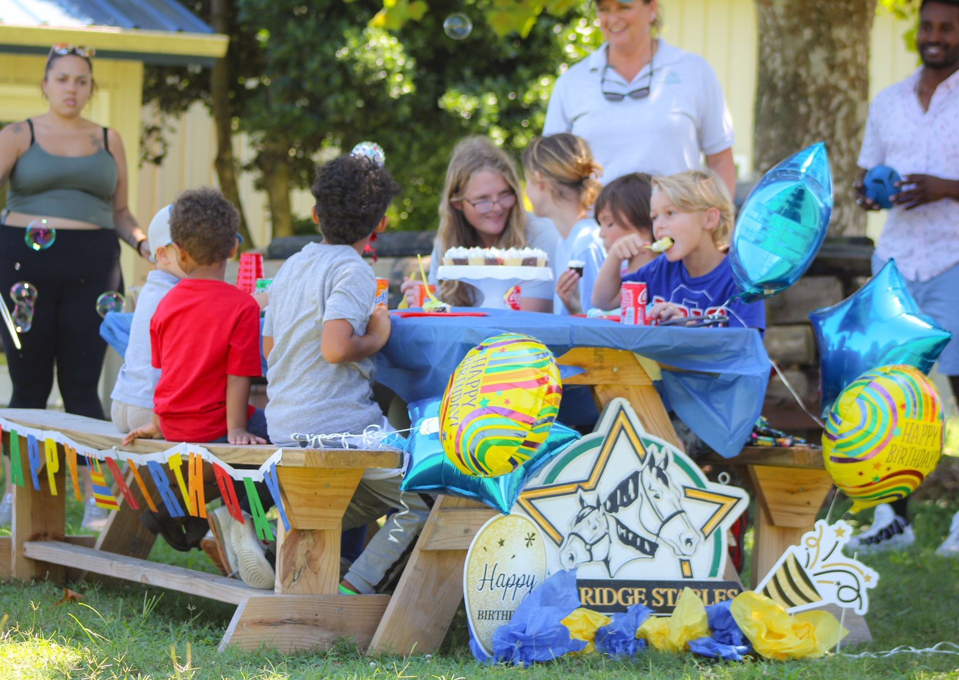 Children seated at a picnic table with cupcakes and balloons at an outdoor birthday party