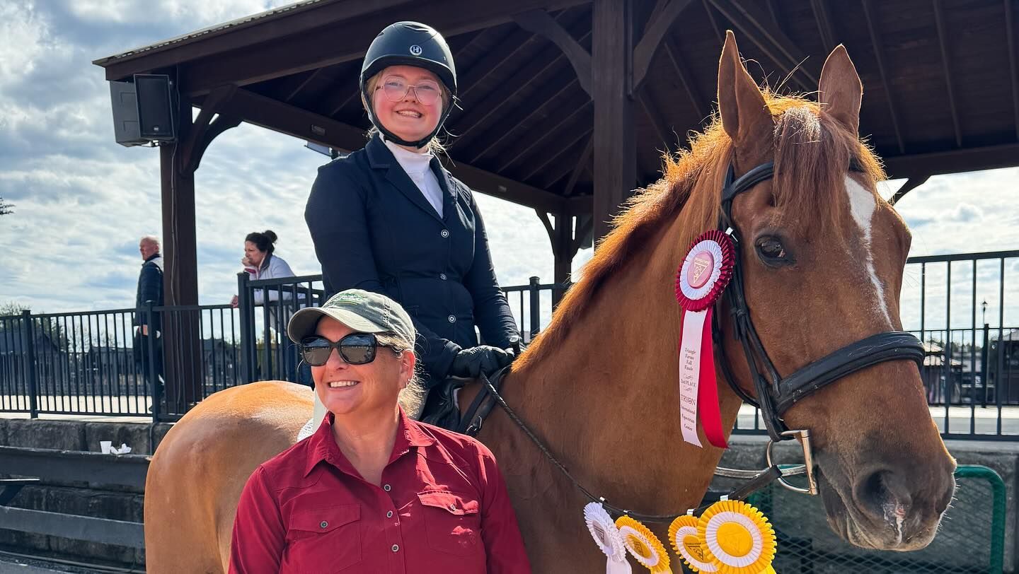 Person on a horse with medals, smiling beside another person in sunglasses at an outdoor stable.