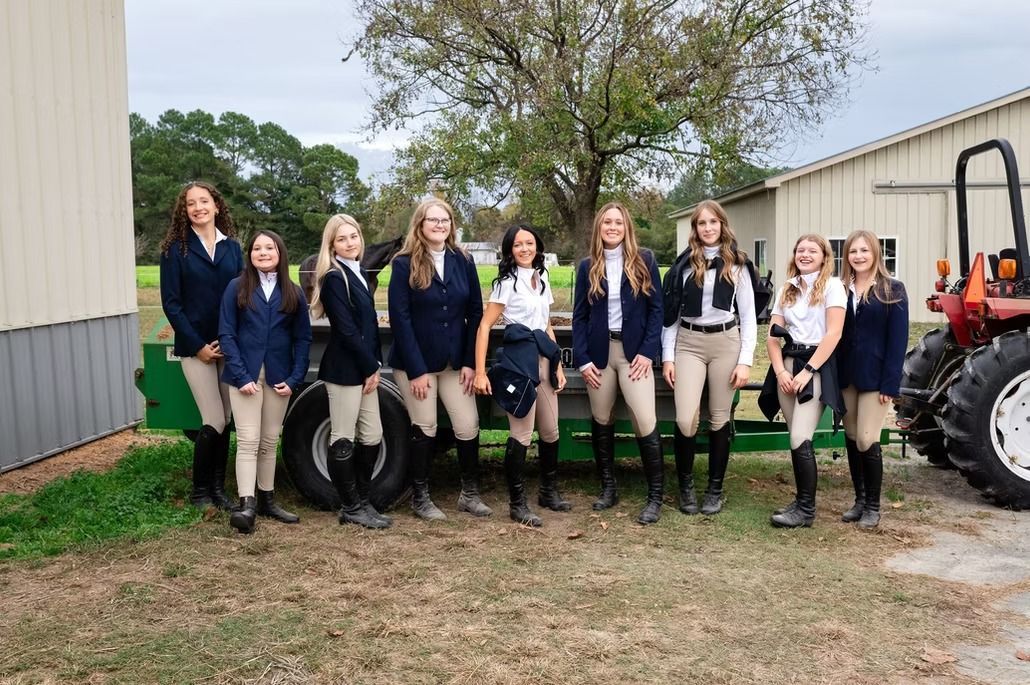 Group of students in navy jackets and beige pants standing outdoors beside farm equipment and a barn