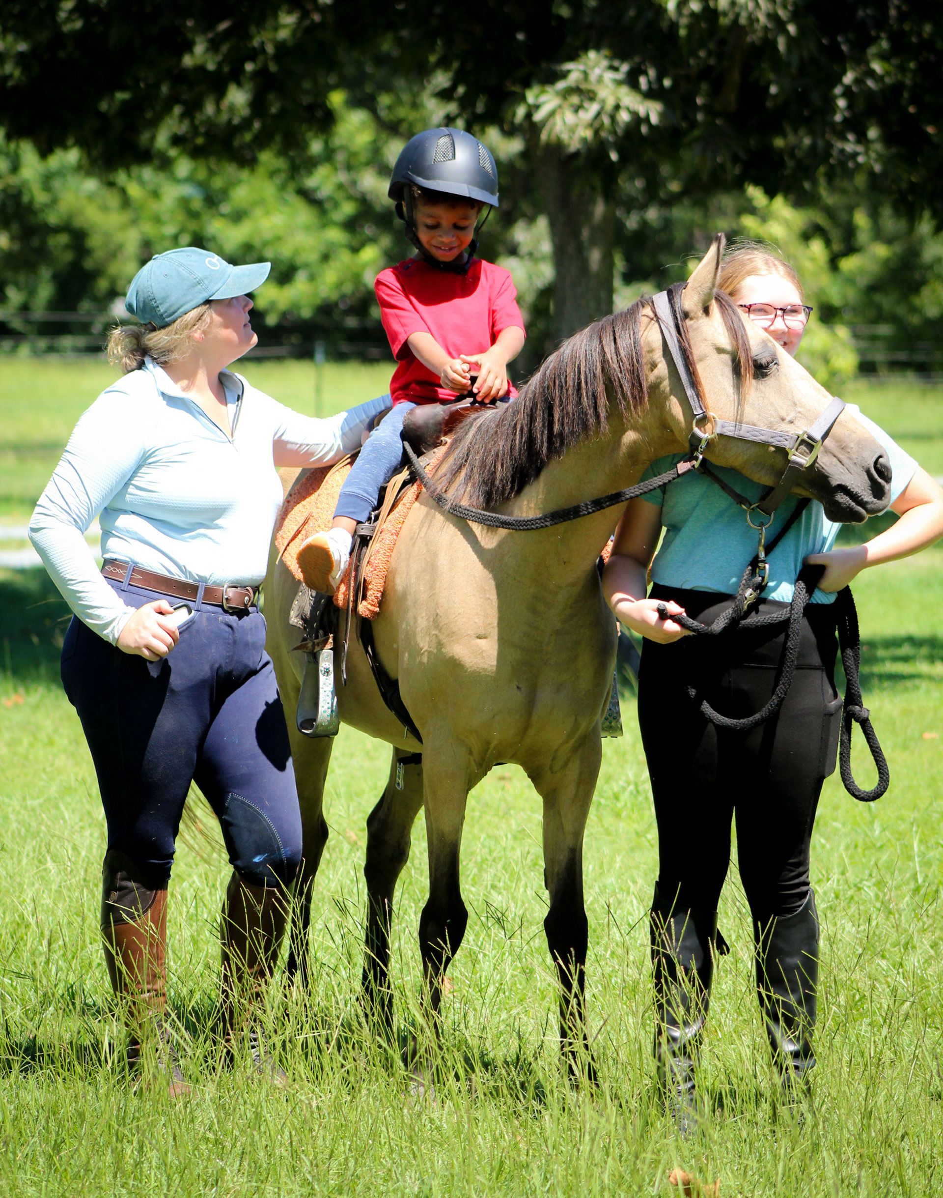 Person riding a pony beside two adults in a grassy field, with trees in the background.