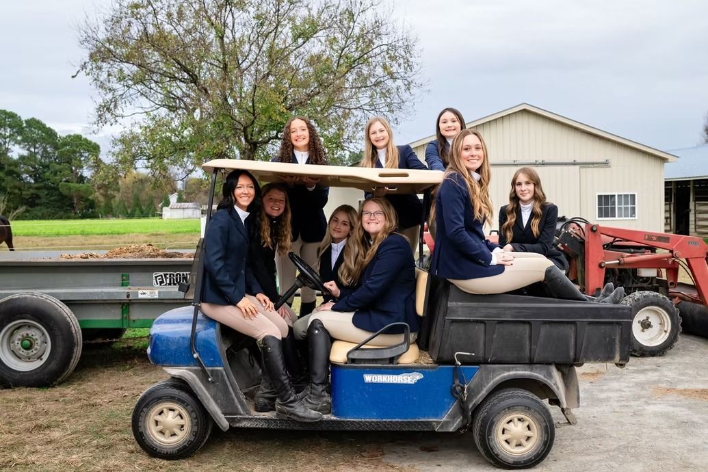 Group seated on a blue golf cart with a trailer, parked outdoors beside a red tractor and barn.