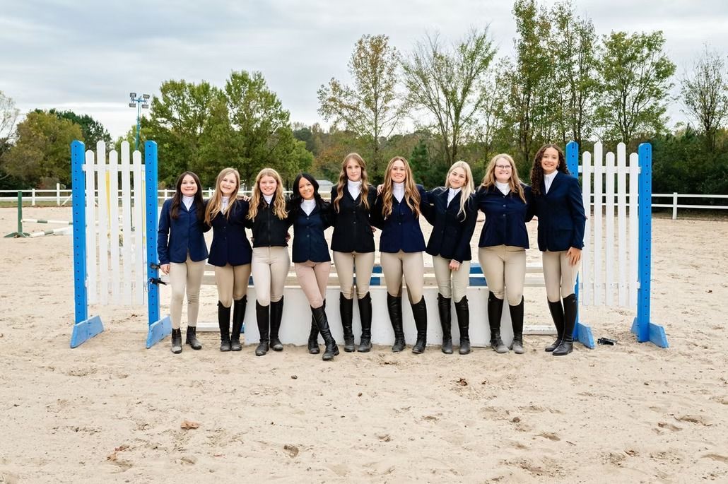 Group in navy riding outfits posing between blue-and-white jumps on a sandy equestrian field.