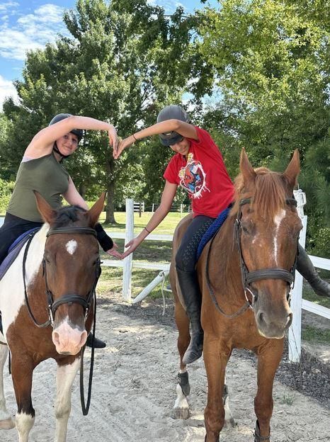 Two riders on horseback make a heart shape with their arms on a sunny trail.