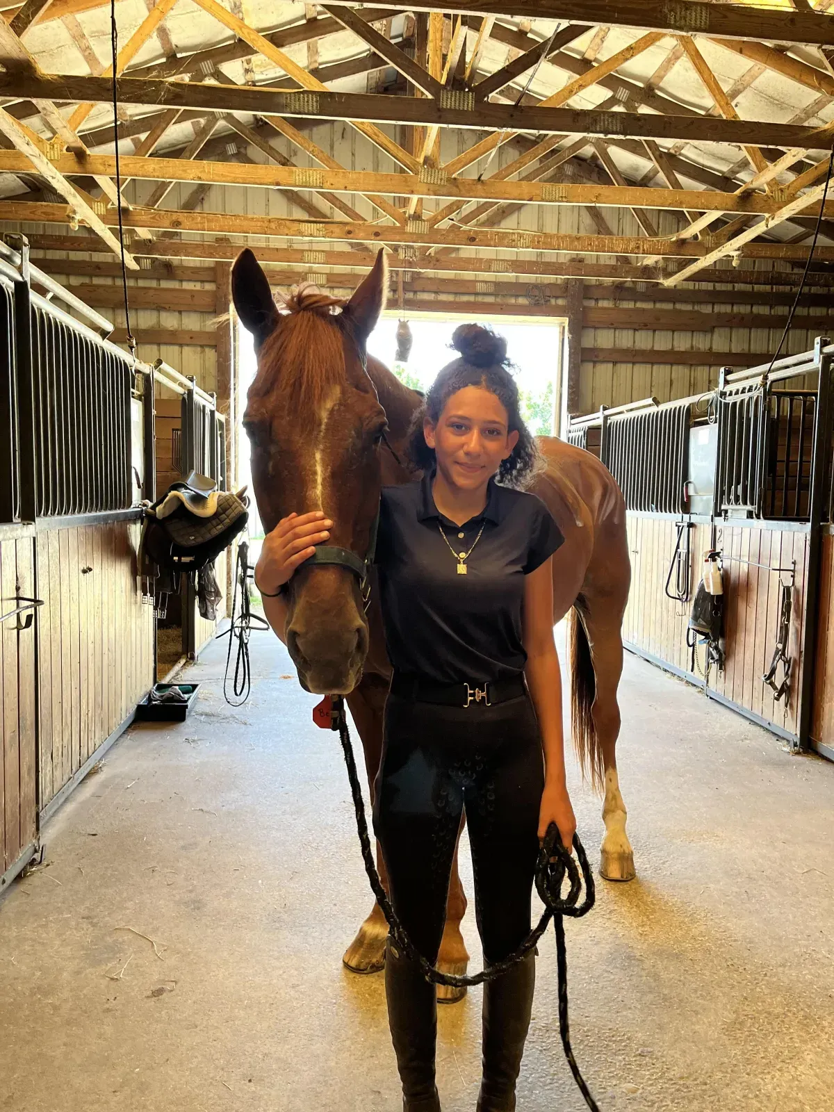 Person leading a brown horse in a stable aisle, with another horse in the background.